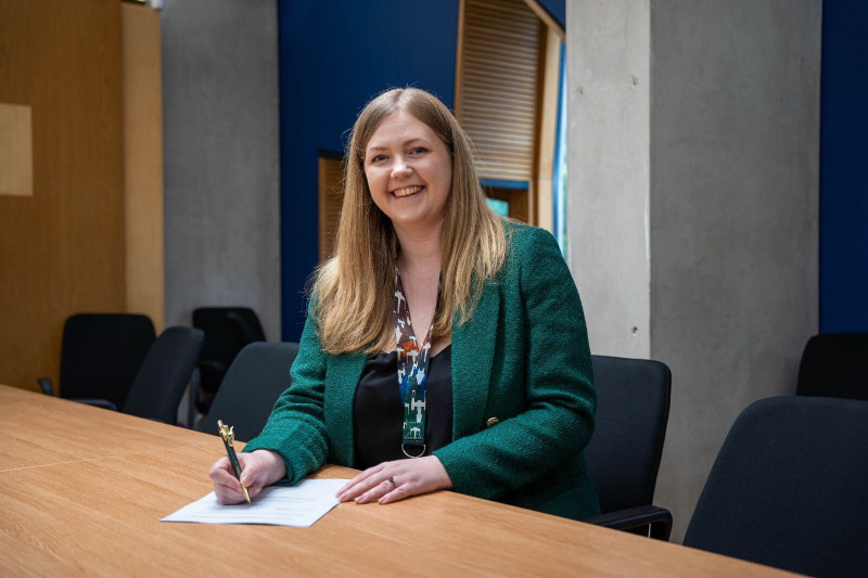 Gillian Mackay wearing green blazer at desk signing Bill